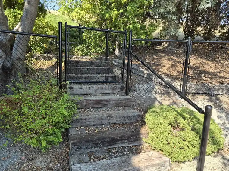Black chain link fence rising beside timber steps on a terraced Yakima yard with retaining walls