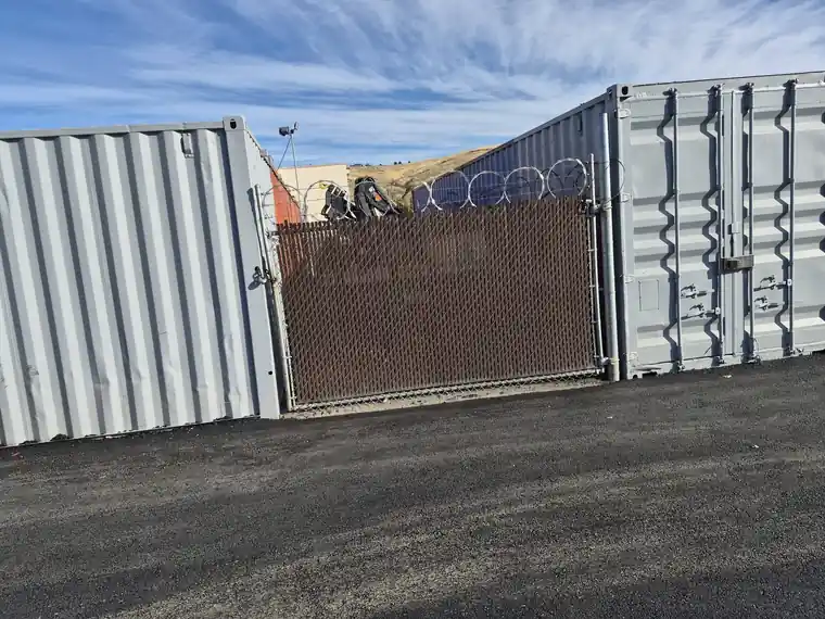 Commercial chain link gate with brown privacy slats and razor wire between shipping containers